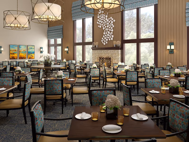 A spacious dining room with multiple tables and chairs arranged neatly. Each table is set with plates, napkins, and glasses, and decorated with small flower arrangements. The room features large windows letting in natural light, a stone fireplace with decorative art above it, and modern hanging light fixtures. The walls have wood paneling and are adorned with paintings and sconces.