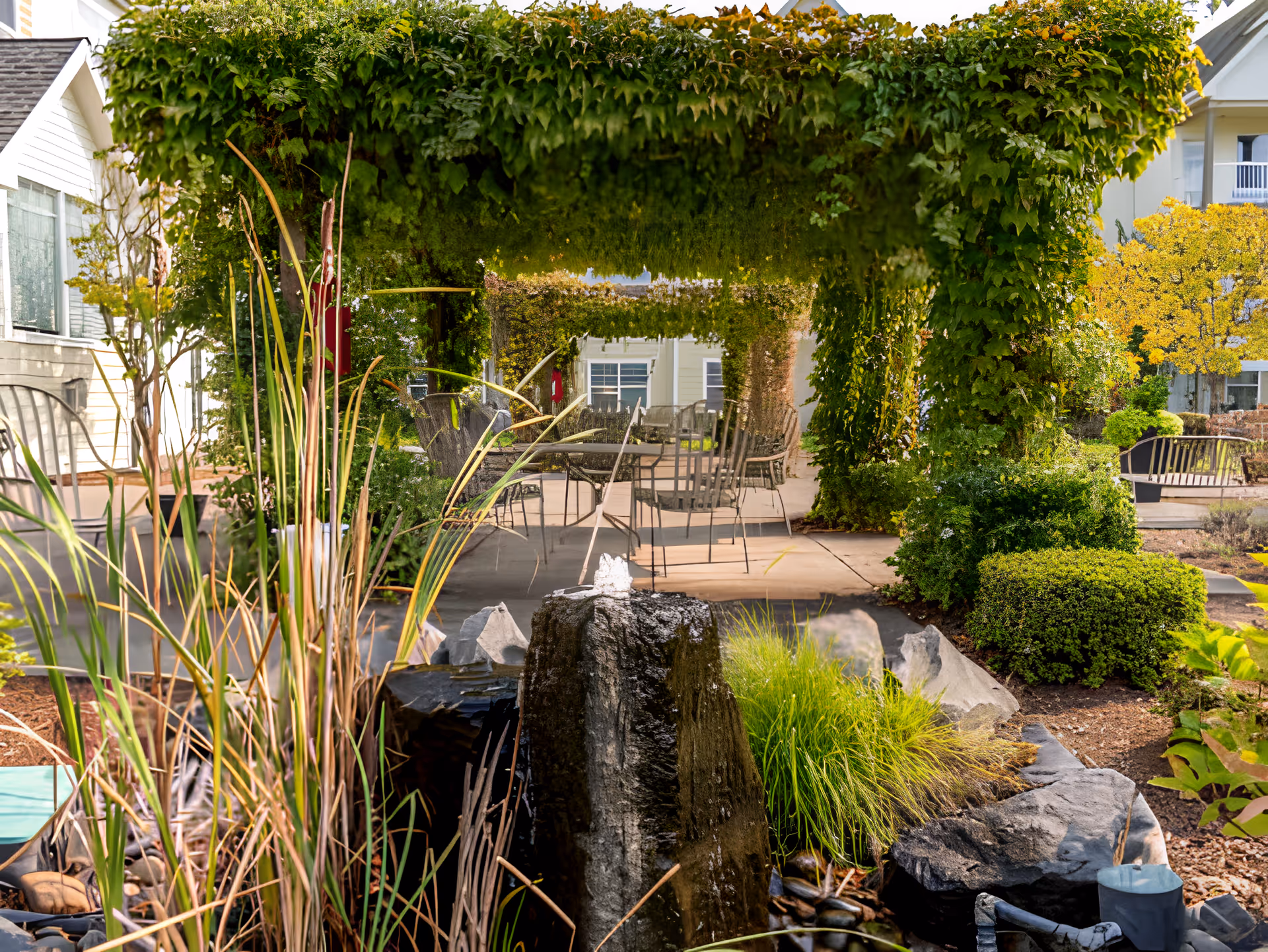 Outdoor garden area at Avamere at Bethany featuring a stone water fountain surrounded by plants and rocks, with a shaded seating area covered by lush green vines and metal chairs and tables in the background.