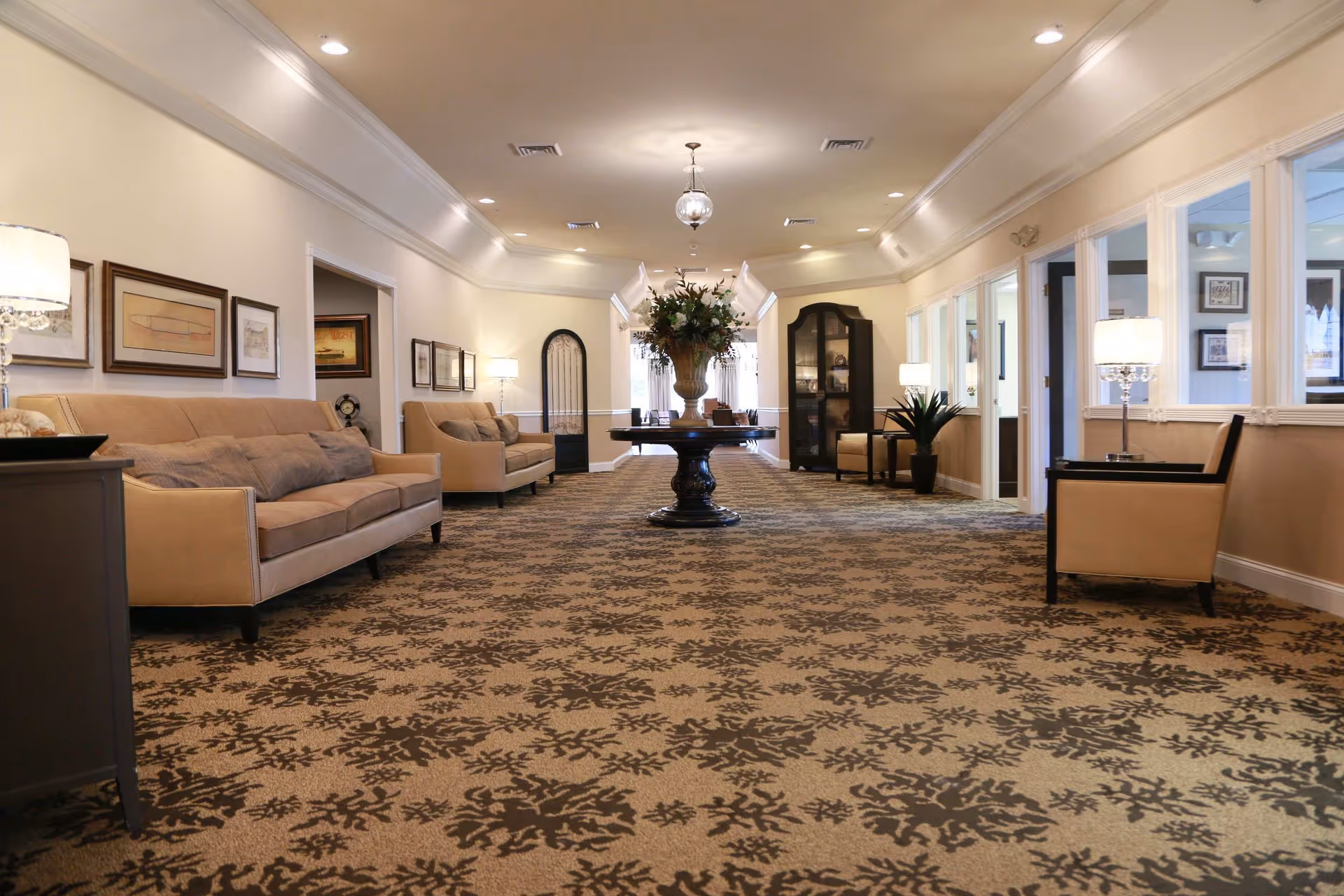 A spacious, well-lit lounge area with beige sofas and armchairs arranged along the walls. The carpet has a decorative floral pattern, and a round table with a large floral arrangement is centered in the room. The walls are adorned with framed artwork, and there are several lamps providing warm lighting. Large windows on the right side allow natural light to enter the space.