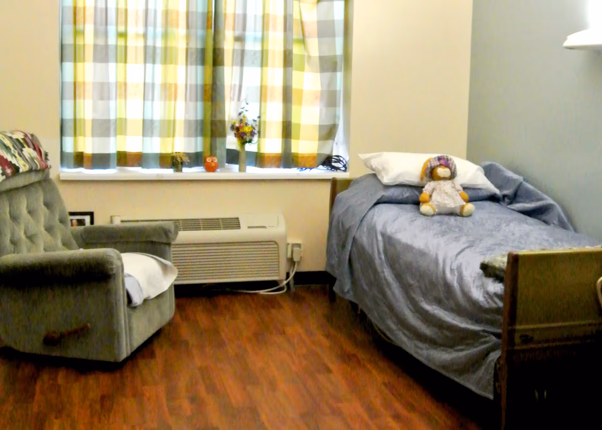 A small bedroom with a single bed covered in a gray blanket and a pillow with a stuffed animal on it. Next to the bed is a green armchair with a white cloth draped over one arm. The room has wooden flooring and a window with checkered curtains. Below the window is a heating or air conditioning unit with some small decorative items on the windowsill.