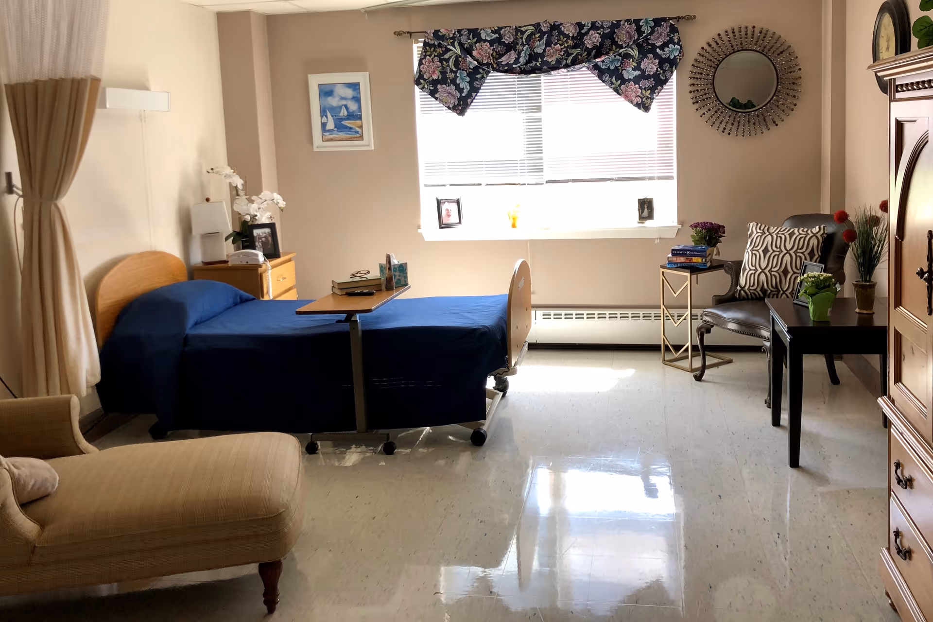 A well-lit room with a single hospital-style bed covered with a blue blanket, a wooden headboard, and an overbed table with books and tissues. To the left, there is a beige chaise lounge and a curtain. On the right side, there is a black armchair with a patterned pillow, a small side table with books and a flower pot, and a larger wooden cabinet. A window with blinds and a floral valance lets in natural light. The walls are decorated with a framed picture and a round mirror.