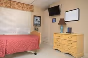 A bedroom in a senior living facility featuring a single bed with a red quilted bedspread, a window with closed blinds and a floral valance, a yellow dresser with a lamp and framed pictures on top, a wall-mounted flat-screen TV, and a whiteboard with the word 'Welcome' written on it.