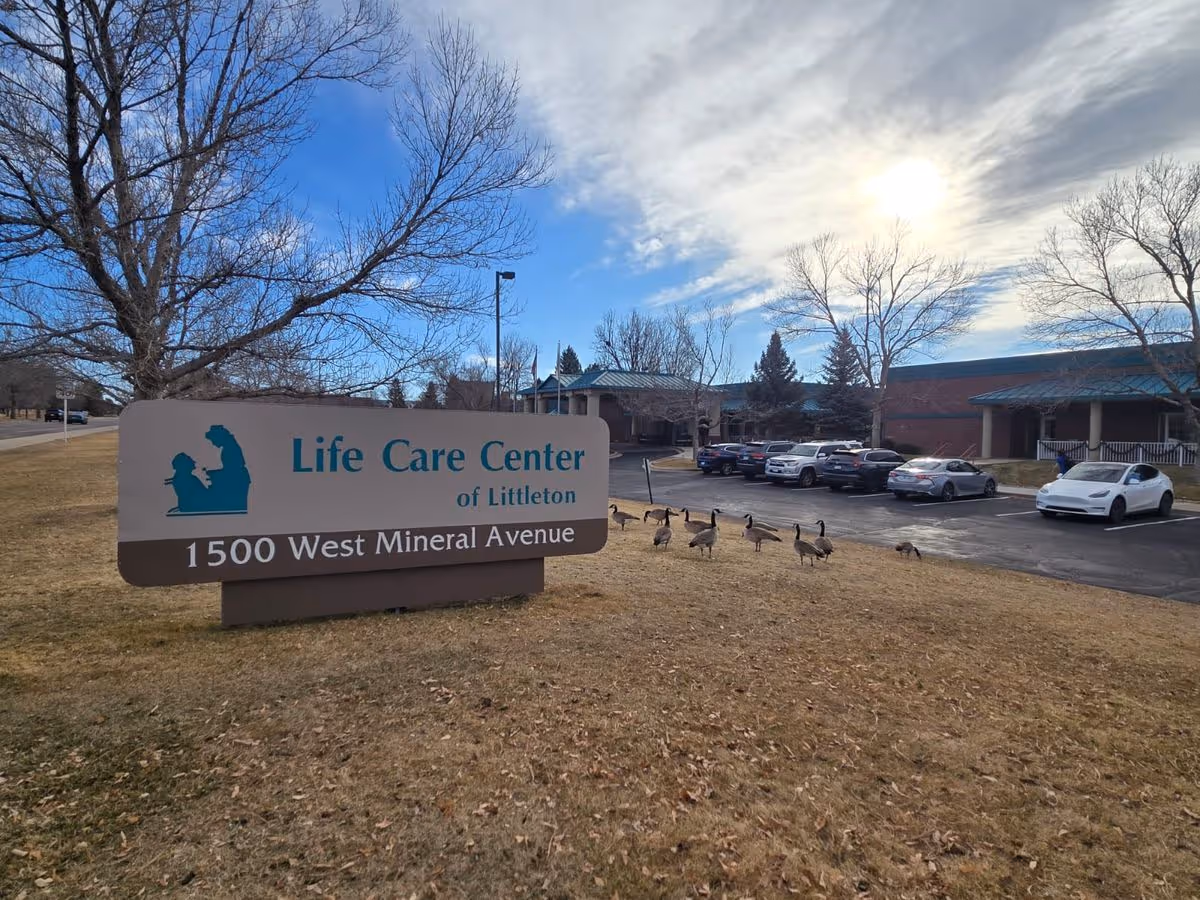 Sign for Life Care Center of Littleton on a grassy lawn with geese, parking lot, and the facility building under a partly cloudy sky.