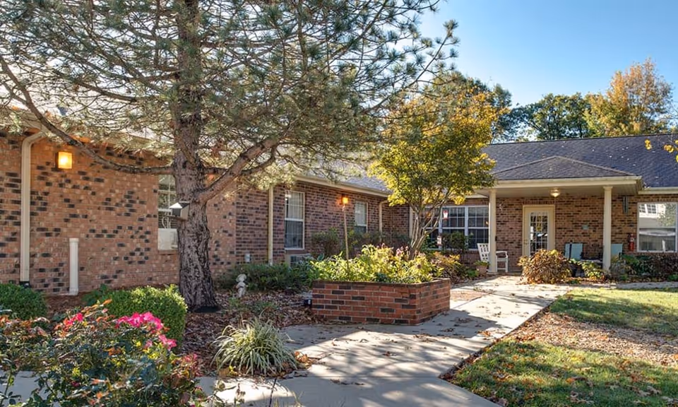Outdoor view of a senior living facility courtyard with a brick building, trees, shrubs, and a paved walkway leading to a covered entrance with chairs.