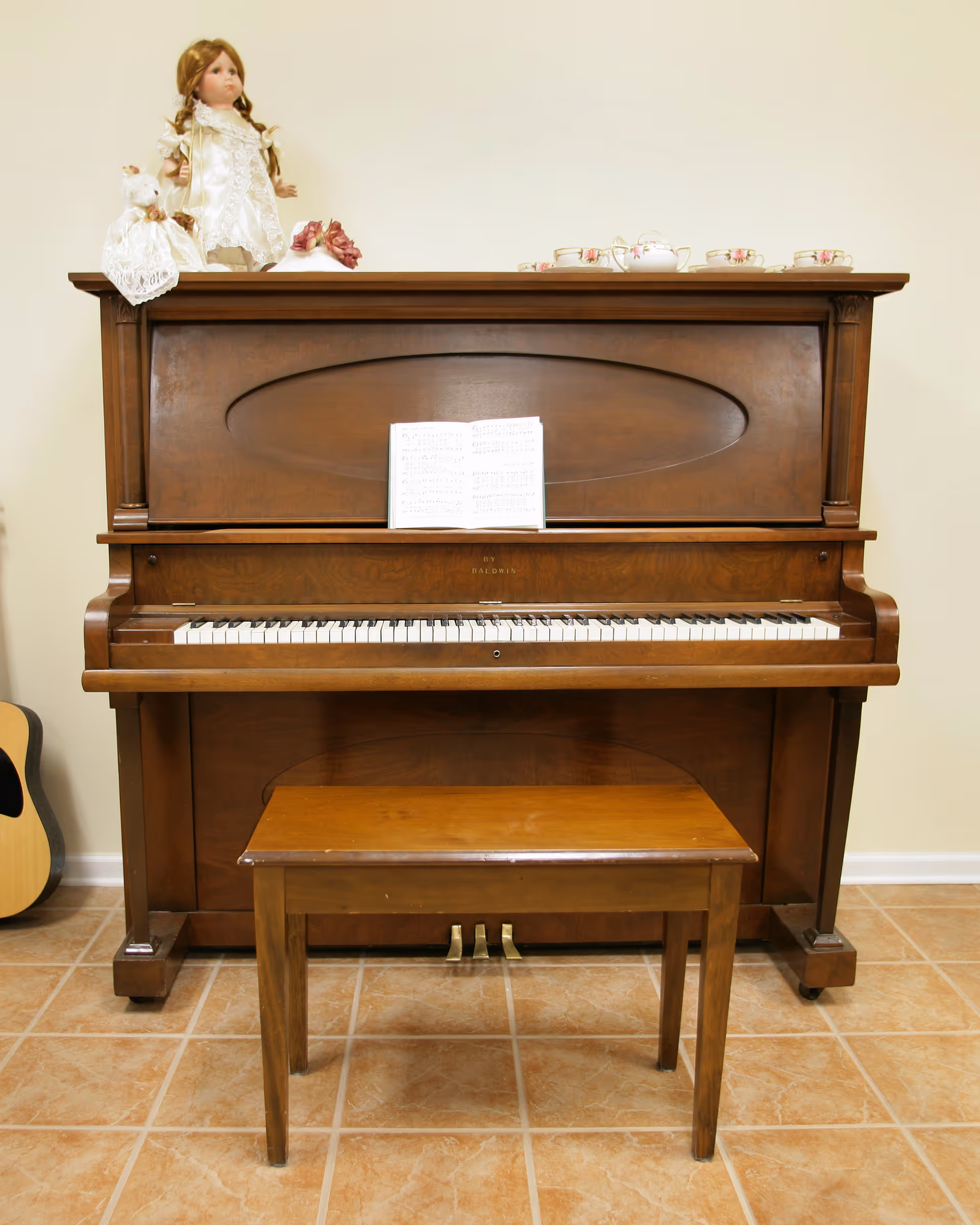 A wooden upright piano with a matching wooden bench in front of it, a sheet of music placed on the piano. On top of the piano are two porcelain dolls dressed in white and a set of floral teacups and a teapot. A guitar is partially visible leaning against the wall to the left.