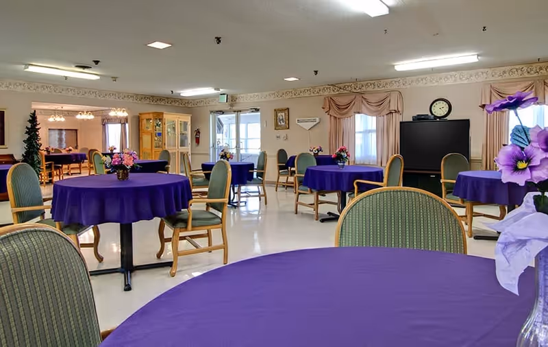 Spacious communal dining room with round tables draped in purple tablecloths, chairs, floral centerpieces, and a TV on the far wall.