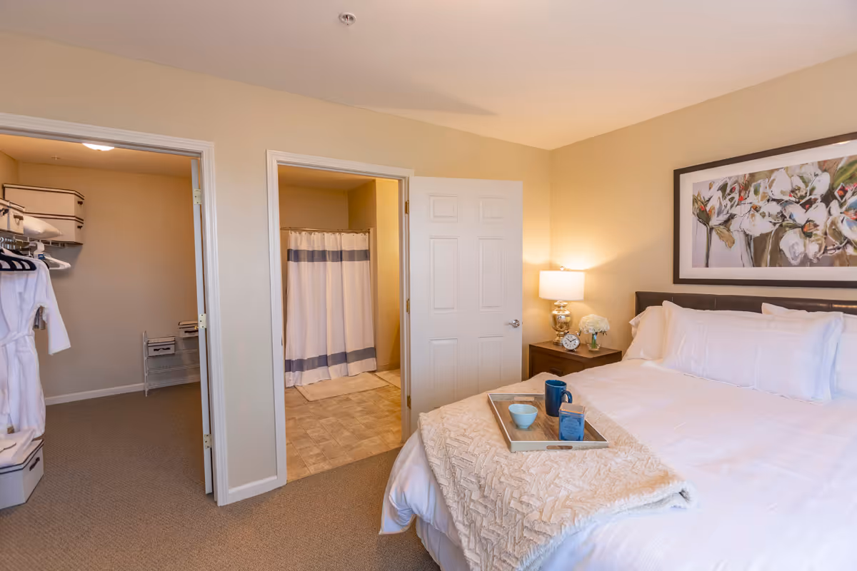 A cozy bedroom with a neatly made bed featuring white linens and a beige throw blanket. A tray with a blue mug, bowl, and a small box is placed on the bed. To the right of the bed is a nightstand with a lamp, a clock, and a vase of white flowers. On the wall above the bed is a large framed floral painting. The room has beige walls and carpeted floors. Two open doors reveal a walk-in closet with hanging clothes and storage boxes on the left, and a bathroom with a shower curtain on the right.