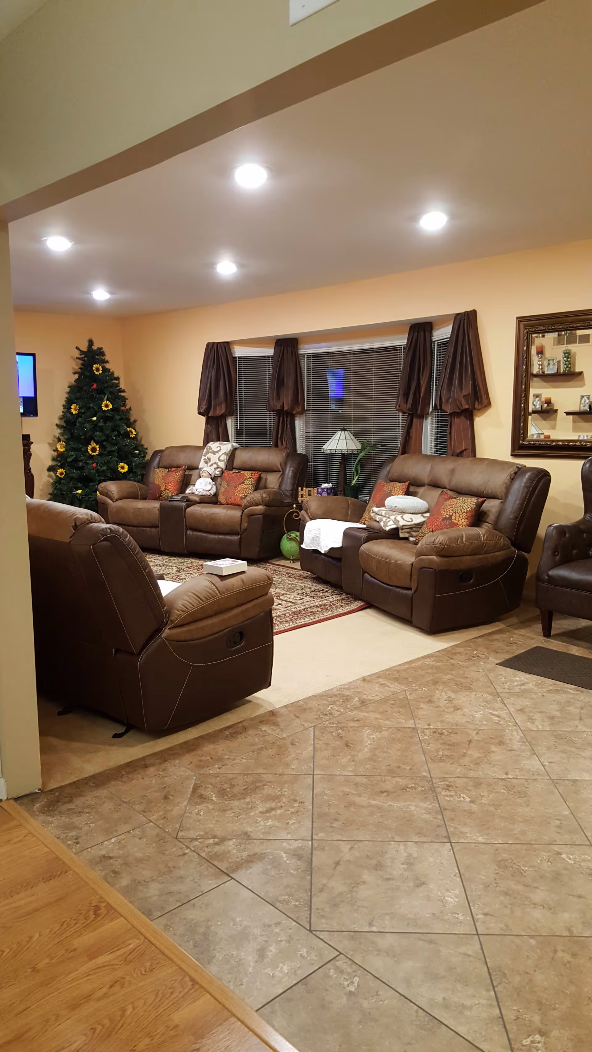 A cozy living room with brown leather recliner sofas arranged around a patterned rug. There is a decorated Christmas tree in the corner, a window with brown curtains, a table lamp, and a wall mirror with shelves displaying decorative items. The floor transitions from tiled to carpeted area.