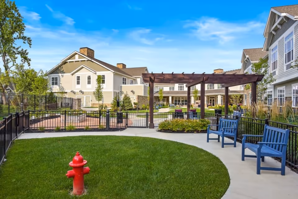 Courtyard of a senior living community with a green lawn and red fire hydrant, a wooden pergola, blue benches, and surrounding multi-story buildings under a blue sky.