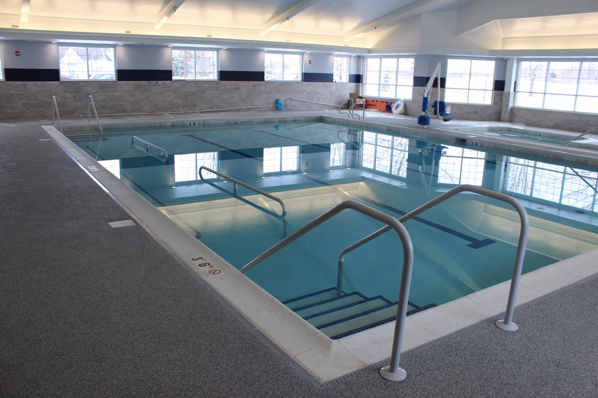 Indoor swimming pool with handrails and steps leading into the water. Large windows along the walls allow natural light to fill the room. The pool area has a non-slip floor and safety equipment is visible in the background.