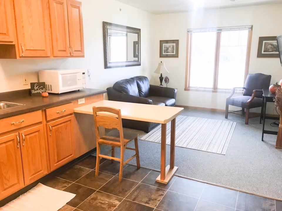 Small open living area with a kitchenette, a table and chair, a leather sofa, an armchair, and a large window.