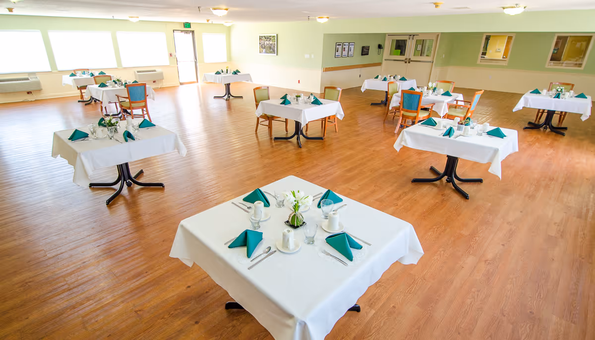 Spacious dining room with several tables set with white tablecloths, green napkins, and place settings on a wood floor.