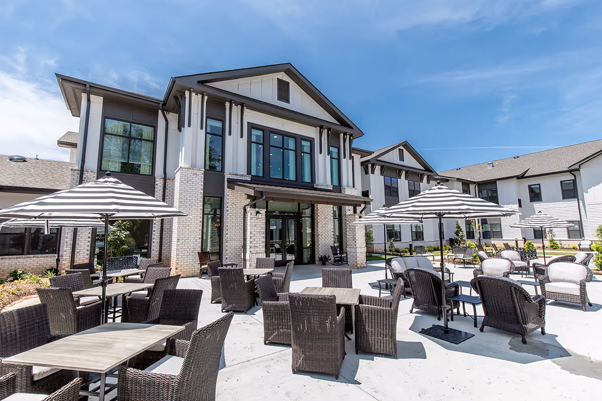 Outdoor patio with tables, wicker chairs and striped umbrellas in front of a modern two-story building under a blue sky.