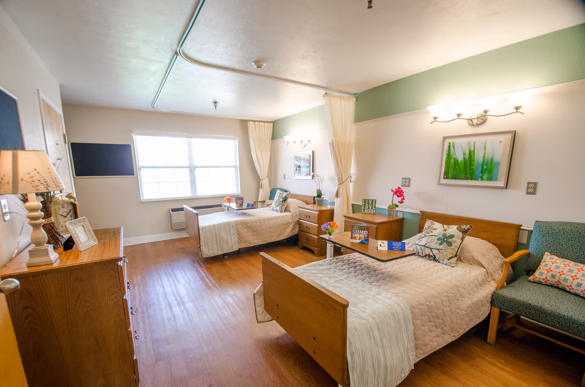 A bright and clean senior living bedroom with two single beds, each with a bedside table and decorative pillows. The room has wooden flooring, a large window letting in natural light, a green accent wall, and a comfortable chair with a patterned cushion. There are framed pictures on the walls and small flower arrangements on the tables.