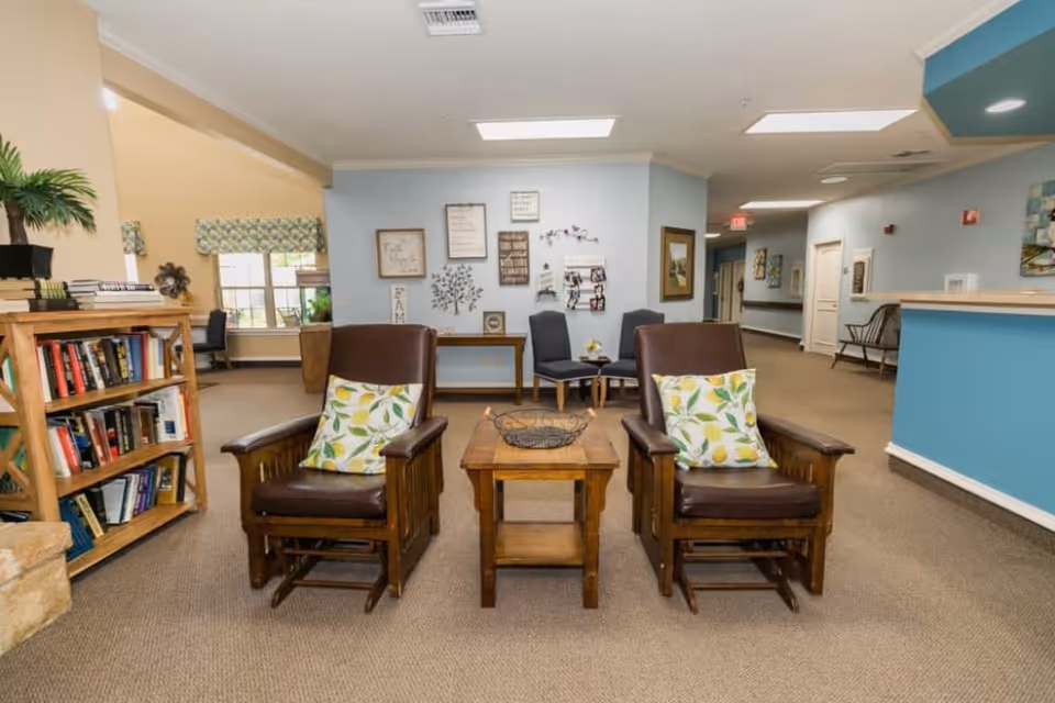 A cozy seating area in a senior living facility with two wooden armchairs featuring brown cushions and lemon-patterned pillows, a wooden side table with a decorative bowl between them, a bookshelf filled with books on the left, and a hallway with additional seating and wall decorations in the background.
