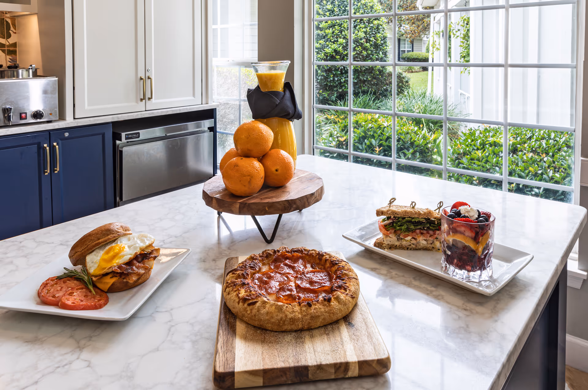 A kitchen countertop with a pepperoni pizza on a wooden cutting board, a breakfast sandwich with egg, bacon, and cheese on a plate with tomato slices, a sandwich with lettuce and tomato on a rectangular plate next to a glass of mixed berries topped with whipped cream. A wooden stand holds a carafe of orange juice and several whole oranges. Large windows in the background show green bushes and a lawn outside.