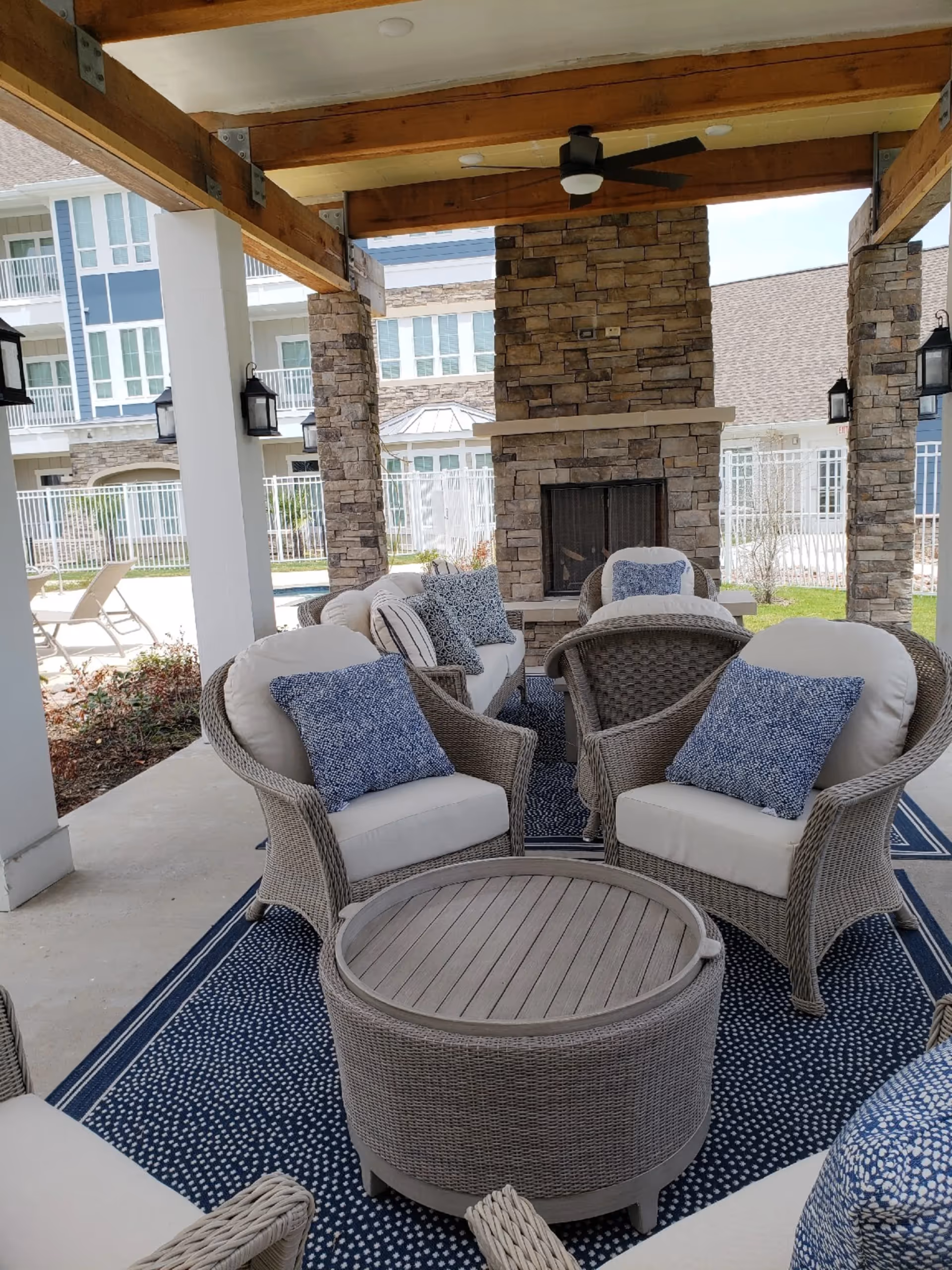 Covered outdoor seating area with wicker chairs and a round wicker table on a blue patterned rug. The seating area is arranged around a stone fireplace with a ceiling fan above. In the background, there are lounge chairs near a pool and a multi-story building with balconies and large windows.