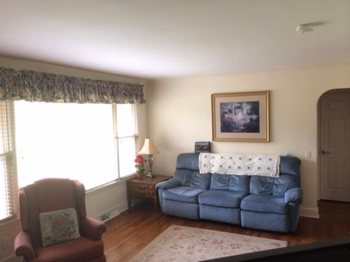A cozy living room with a large window covered by floral valances, a blue three-seat recliner sofa with a white crocheted throw draped over the back, a wooden side table with a lamp and a small potted plant, a framed picture hanging on the wall, and a maroon armchair with a decorative pillow. The room has wooden flooring and light-colored walls.
