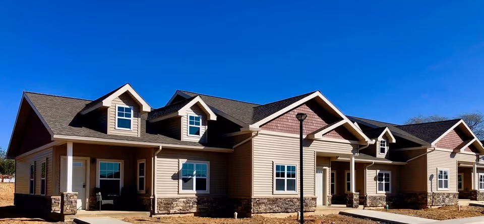 Exterior view of a single-story senior living community building with beige siding, stone accents, multiple windows, and a clear blue sky in the background.