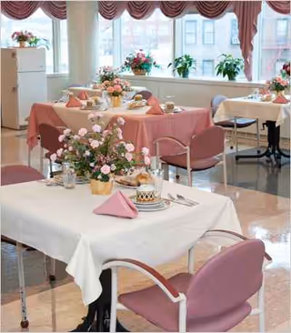 Sunlit dining room with pink tablecloths, flower centerpieces, and pastel chairs.