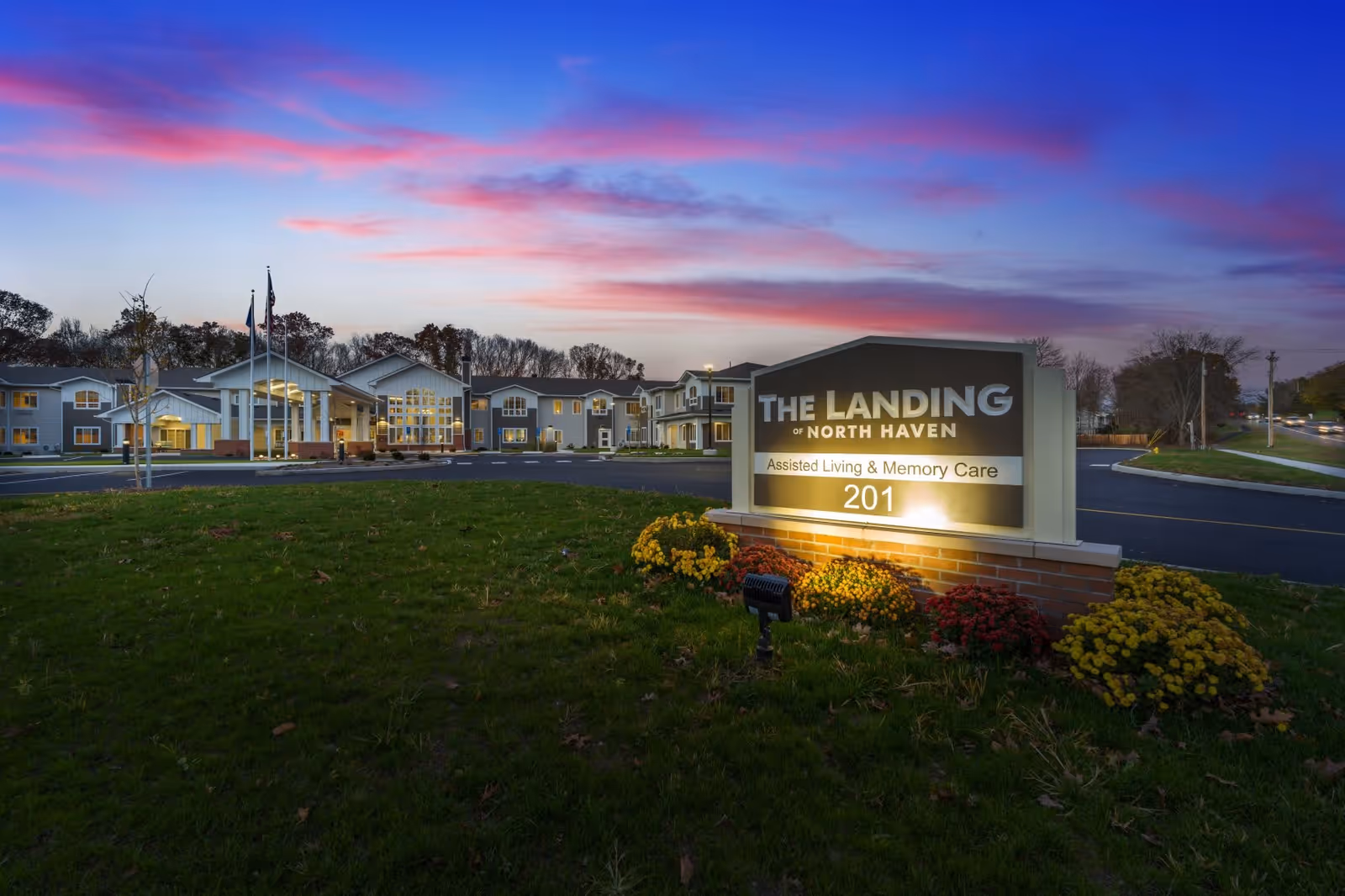 Exterior view of The Landing of North Haven assisted living and memory care facility at dusk, with a well-lit sign in the foreground and the building in the background under a colorful sky.