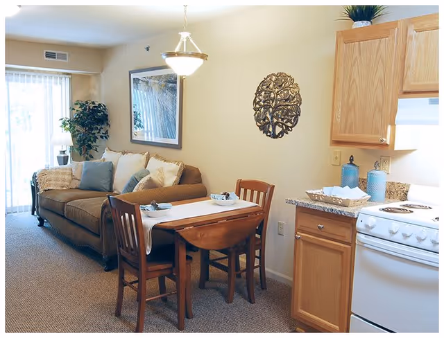 A cozy living and dining area in a retirement facility featuring a brown sofa with cushions, a small wooden dining table with two chairs, a kitchen area with wooden cabinets, a stove, and decorative items including a wall art piece and a potted plant near a sliding glass door.