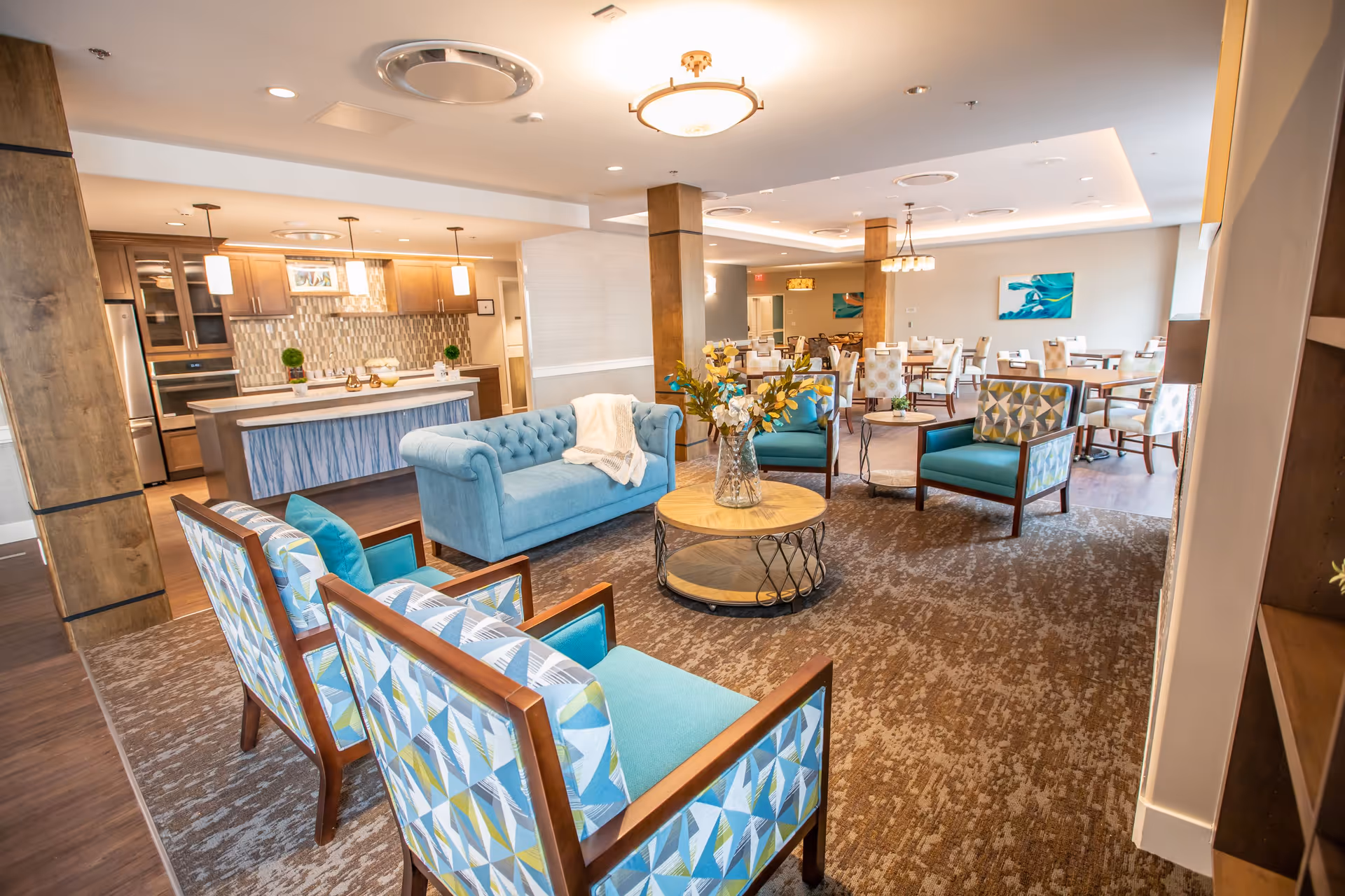 A spacious and well-lit common area in a senior living facility featuring a seating arrangement with a blue tufted sofa, patterned armchairs, and a round wooden coffee table with a vase of flowers. In the background, there is a dining area with multiple tables and chairs, and a kitchen area with wooden cabinets and pendant lighting.