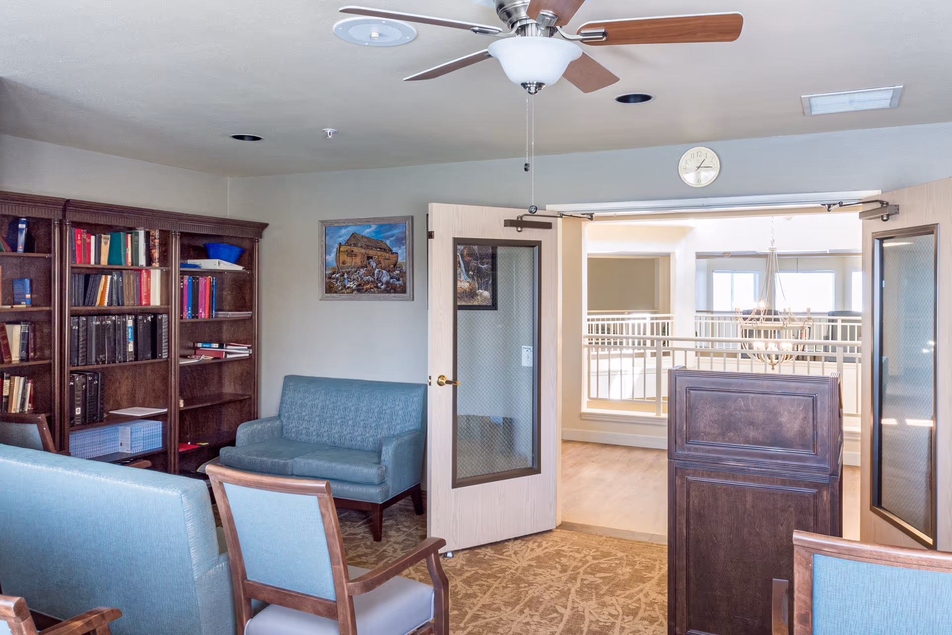 Interior view of a senior living facility room with blue upholstered chairs and a loveseat, a wooden bookshelf filled with books, a ceiling fan, and double doors opening to a bright hallway with a chandelier and railing.