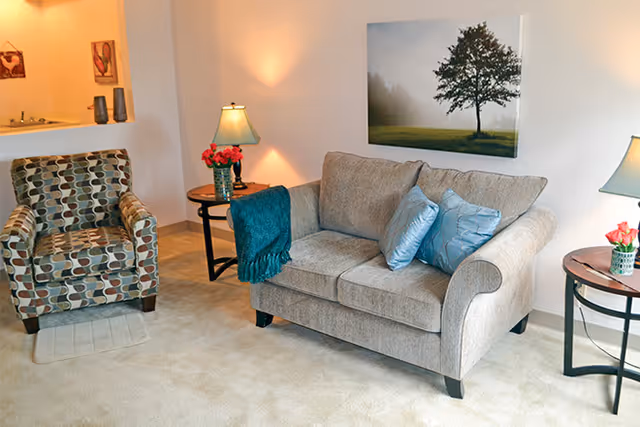 A cozy living room area with a beige loveseat adorned with two blue pillows and a teal throw blanket. To the left is a patterned armchair with geometric shapes. There are two round wooden side tables, each with a lamp and a small vase of flowers. A large framed picture of a tree in a misty field hangs on the wall above the loveseat. The room has soft lighting and beige carpeting.