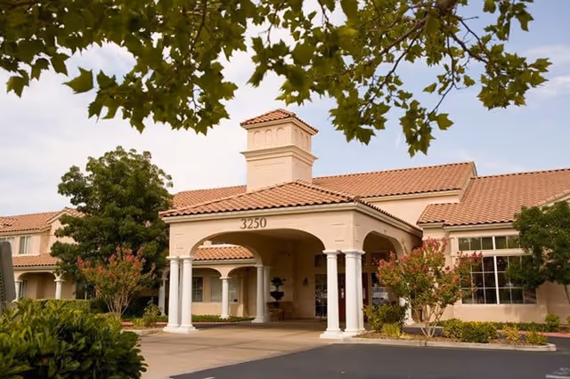 Exterior view of Oakmont of the Lakes senior living facility showing the main entrance with a covered portico supported by white columns, surrounded by trees and landscaping under a partly cloudy sky.