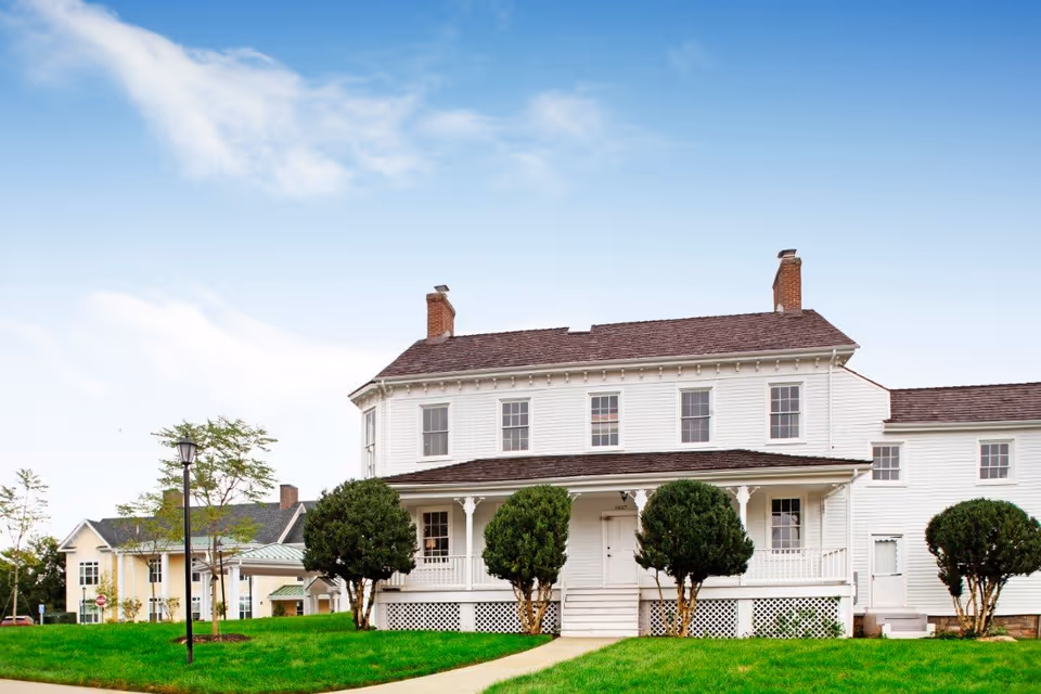 White two-story colonial-style building with a covered front porch, trimmed shrubs and a green lawn under a blue sky.