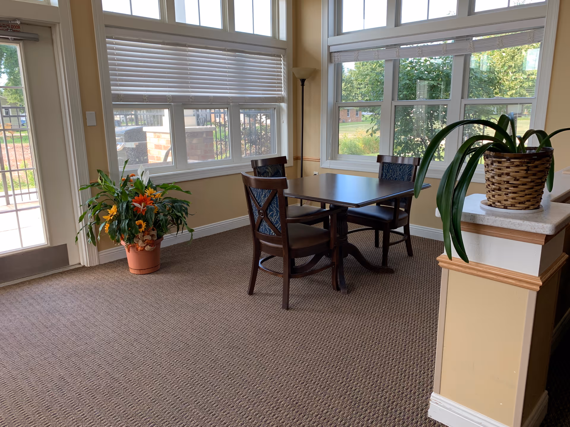 A small seating area with a dark wooden table and four matching chairs with blue patterned cushions, situated near large windows with white blinds. There is a potted plant with colorful flowers on the floor near a glass door, and another green plant in a woven basket on a half wall. The room has beige walls and carpeted flooring.