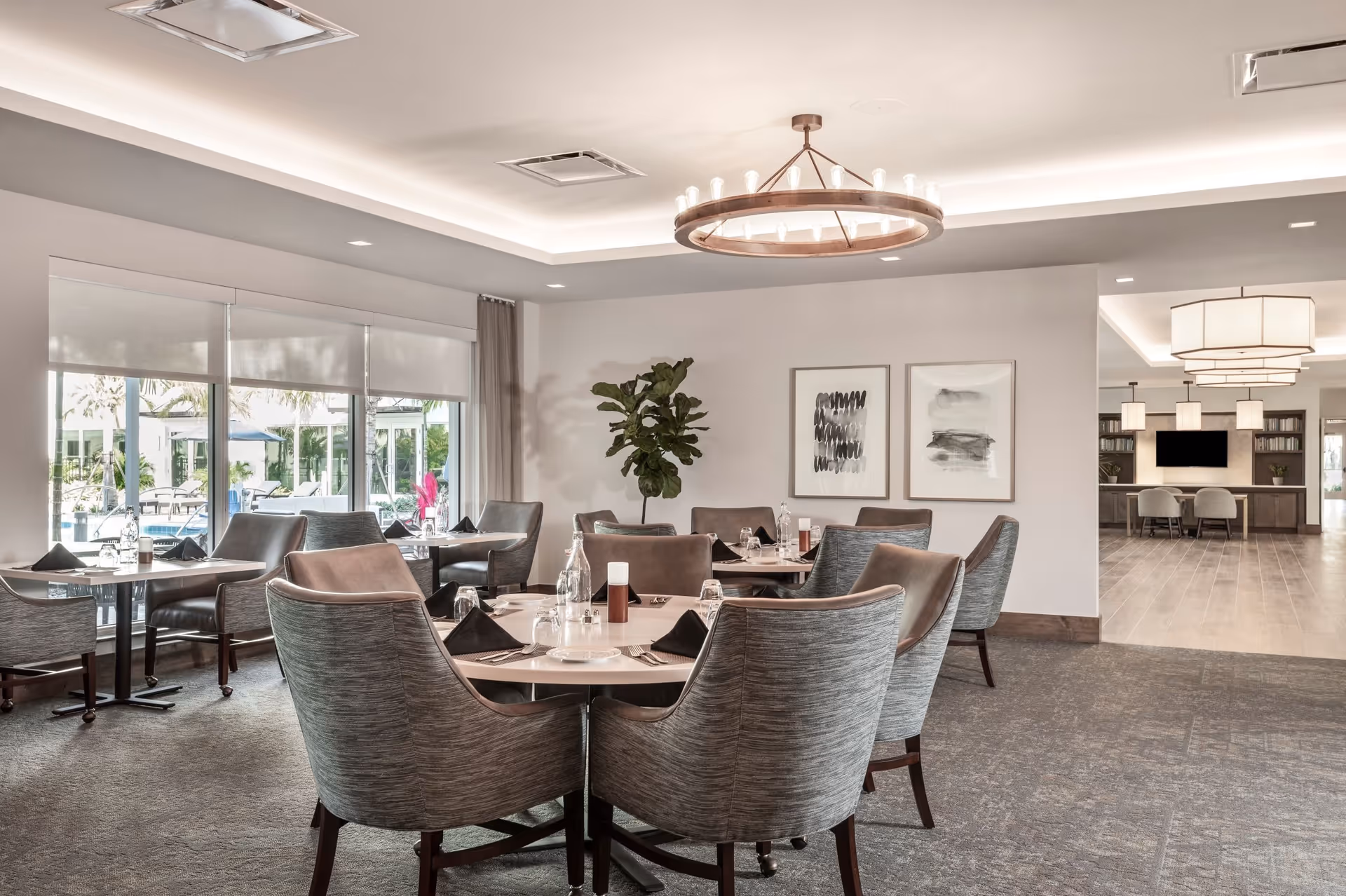 A modern dining room with round tables set with plates, glasses, and black napkins. Comfortable gray upholstered chairs surround the tables. Large windows on the left side provide a view of an outdoor patio area with lounge chairs and umbrellas. The room features neutral tones, contemporary light fixtures, and framed abstract artwork on the walls.