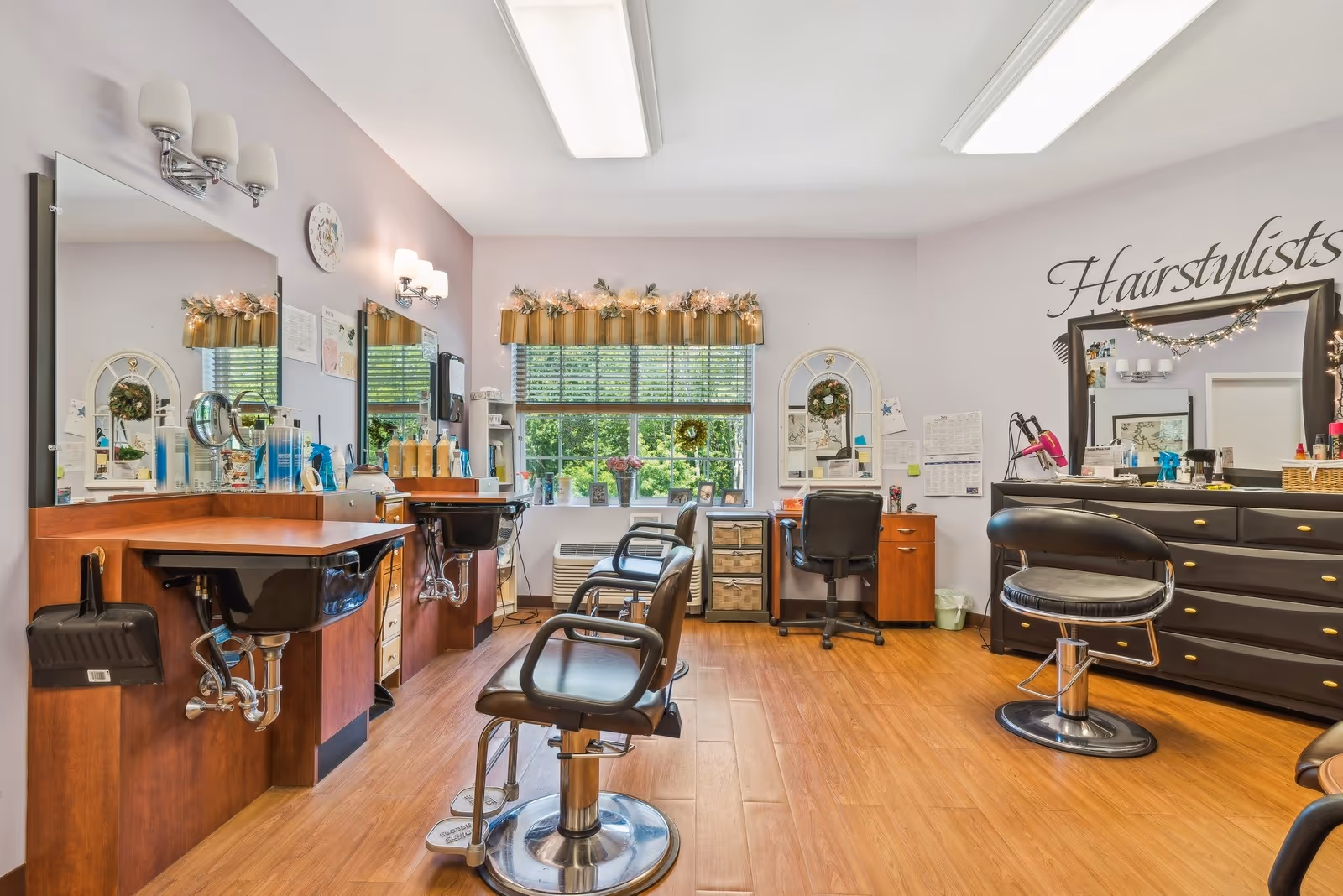 Interior view of a hairstyling salon with multiple styling chairs, mirrors, and sinks. The room has wooden flooring, a window with blinds and decorative valance, and various hair care products on counters. The word 'Hairstylists' is written on the wall above a large mirror and dresser.