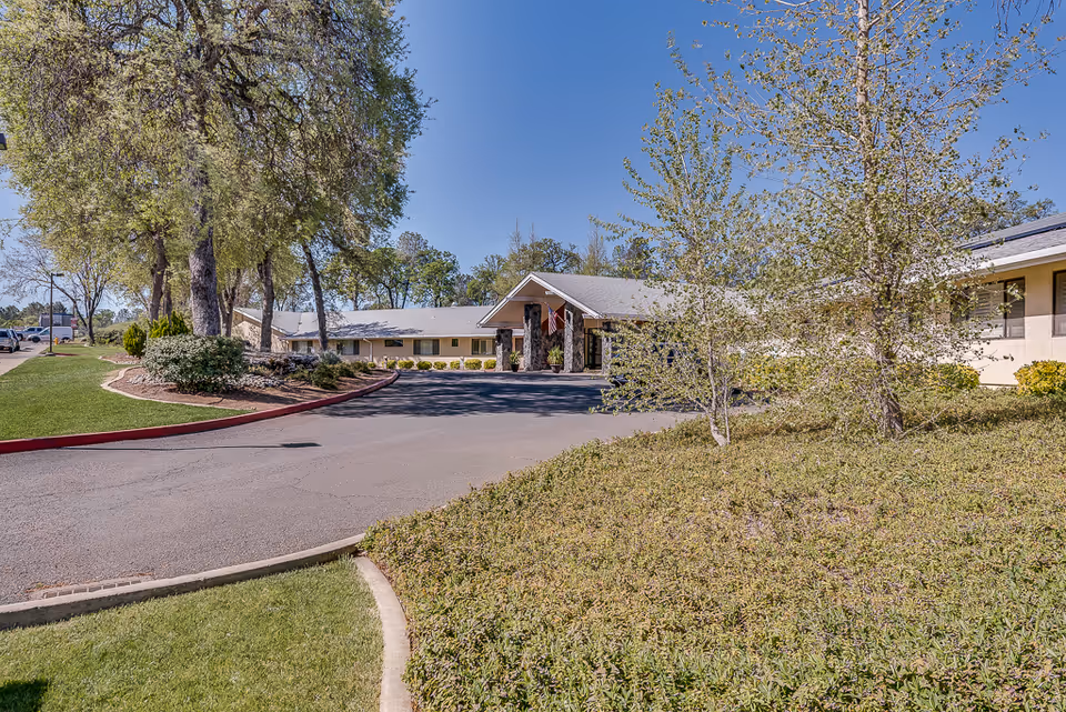 Single-story nursing center front entrance with a circular driveway, flag, trees, and landscaped grounds under a clear blue sky.