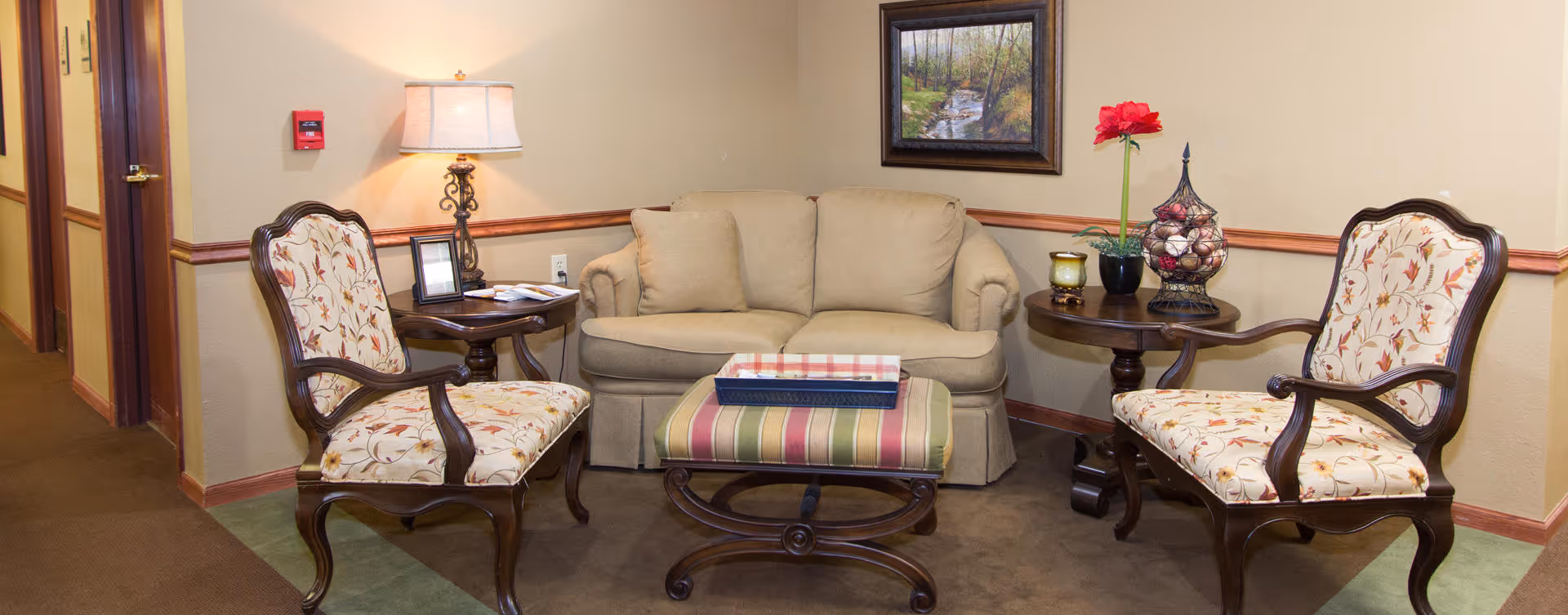 A cozy sitting area in a senior living facility with a beige loveseat, two floral upholstered wooden armchairs, a striped cushioned ottoman, and two wooden side tables. One side table has a lamp and picture frame, while the other has a decorative flower arrangement and a candle. A framed landscape painting hangs on the wall above the loveseat.