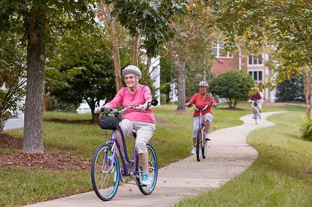 Three elderly women riding bicycles on a winding paved path through a green, tree-lined outdoor area with grass and bushes, near a building in the background.