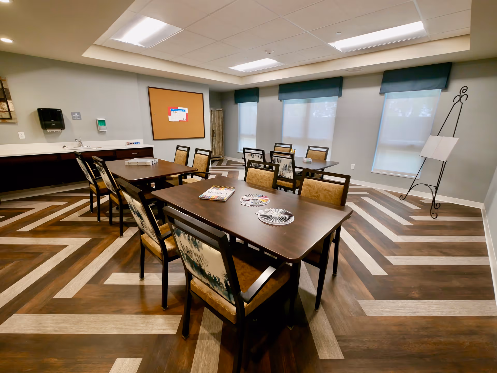 A well-lit room with two rectangular tables surrounded by chairs with patterned upholstery. The floor has a distinctive chevron pattern in dark and light wood tones. The walls are painted light gray with three windows covered by blue valances. There is a cork bulletin board on one wall, a small sink with a paper towel dispenser and soap dispenser, and a decorative easel stand in the corner.