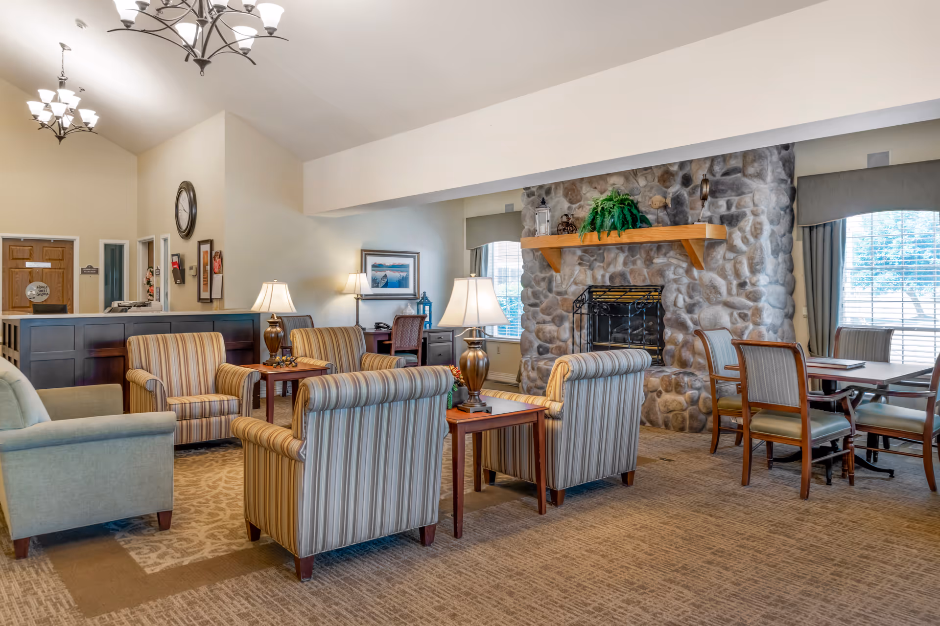 A cozy living room area in a senior living facility featuring several striped armchairs and a green armchair arranged around wooden side tables with lamps. There is a large stone fireplace with a wooden mantle decorated with a green plant and lanterns. In the background, there is a reception desk, a wall clock, framed artwork, and a table with chairs near windows with blinds and curtains.