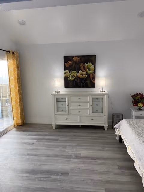Bright bedroom interior with a white dresser topped by two lamps and floral artwork, a partial bed on the right, sliding glass door with yellow curtains on the left, and gray wood flooring.