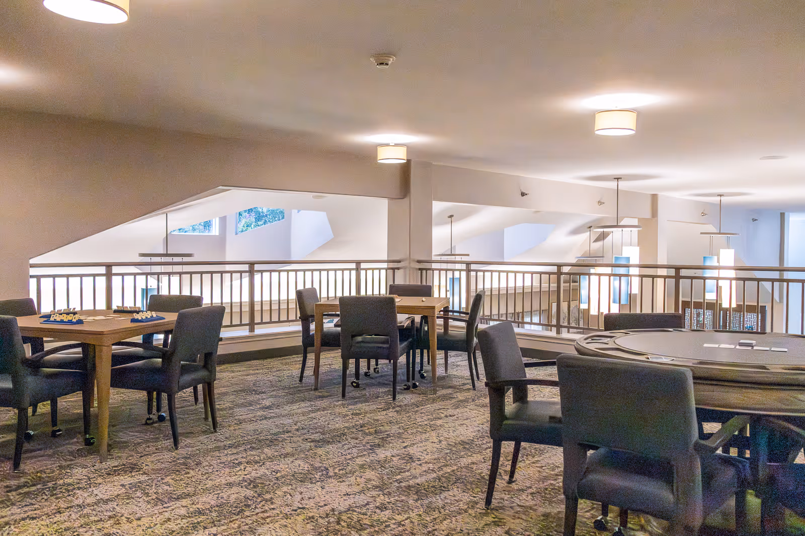 Interior view of a common area in a senior living facility with multiple tables and chairs arranged for social or recreational activities. The space has carpeted flooring, modern lighting fixtures, and a railing overlooking a lower level.