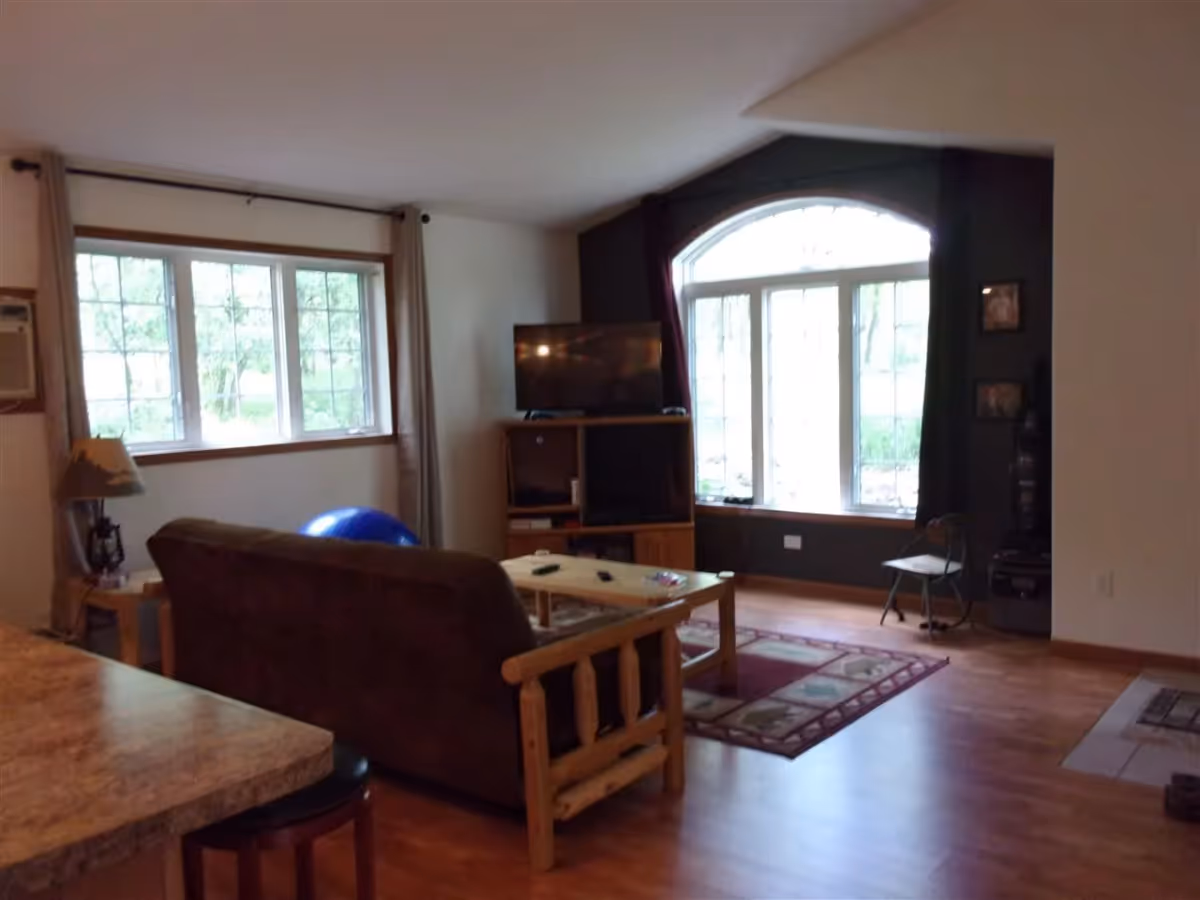 A cozy living room with wooden flooring, a brown couch facing a wooden coffee table, a TV on a wooden stand, large windows with curtains allowing natural light, and a small chair near the window.