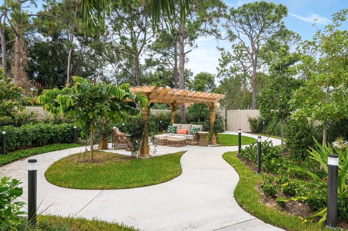 A landscaped outdoor garden area with a curved concrete pathway leading to a wooden pergola with cushioned seating and a small table. The area is surrounded by green grass, various plants, and trees, with a white fence in the background.