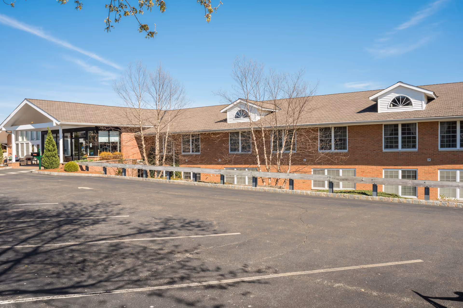 Front exterior of a one-story brick assisted living building with a covered entrance, parking lot, and bare trees under a blue sky.