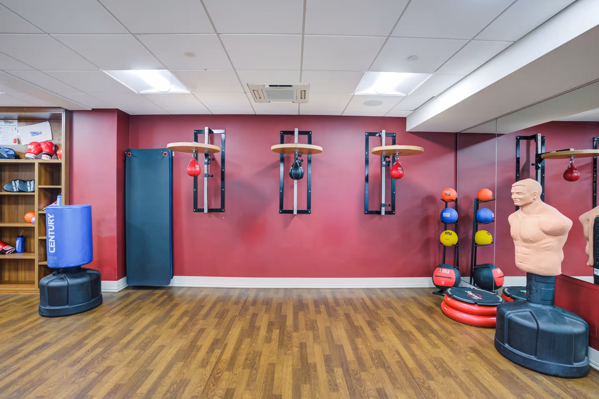 An indoor exercise room with wooden flooring and maroon walls. The room features three mounted speed bags, a blue freestanding punching bag, a black padded mat leaning against the wall, a rack holding various colored medicine balls, and a torso-shaped punching dummy. A large mirror covers the right wall.