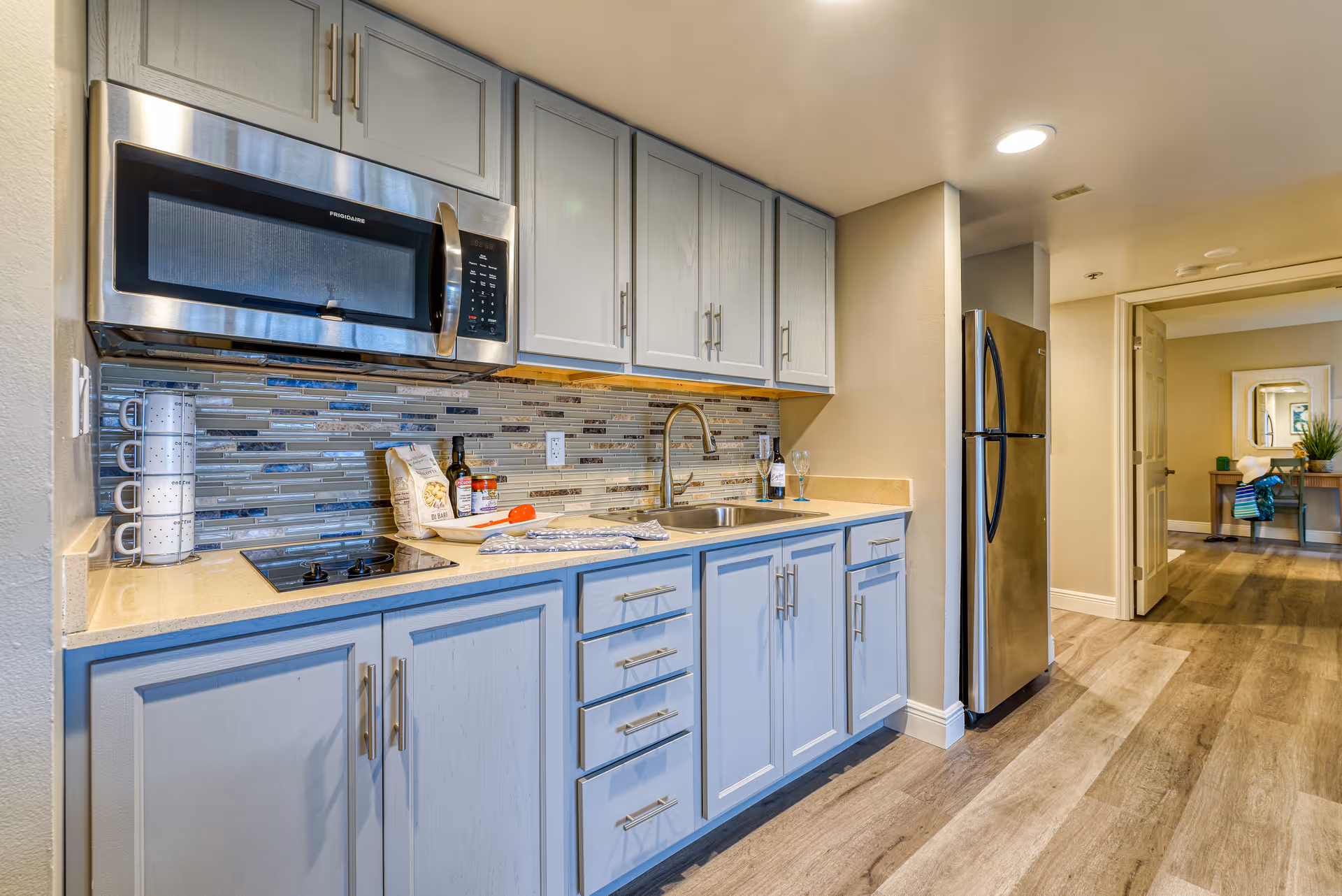 Modern kitchenette with gray cabinets, stainless-steel microwave and refrigerator, sink, and tile backsplash in a senior living apartment.