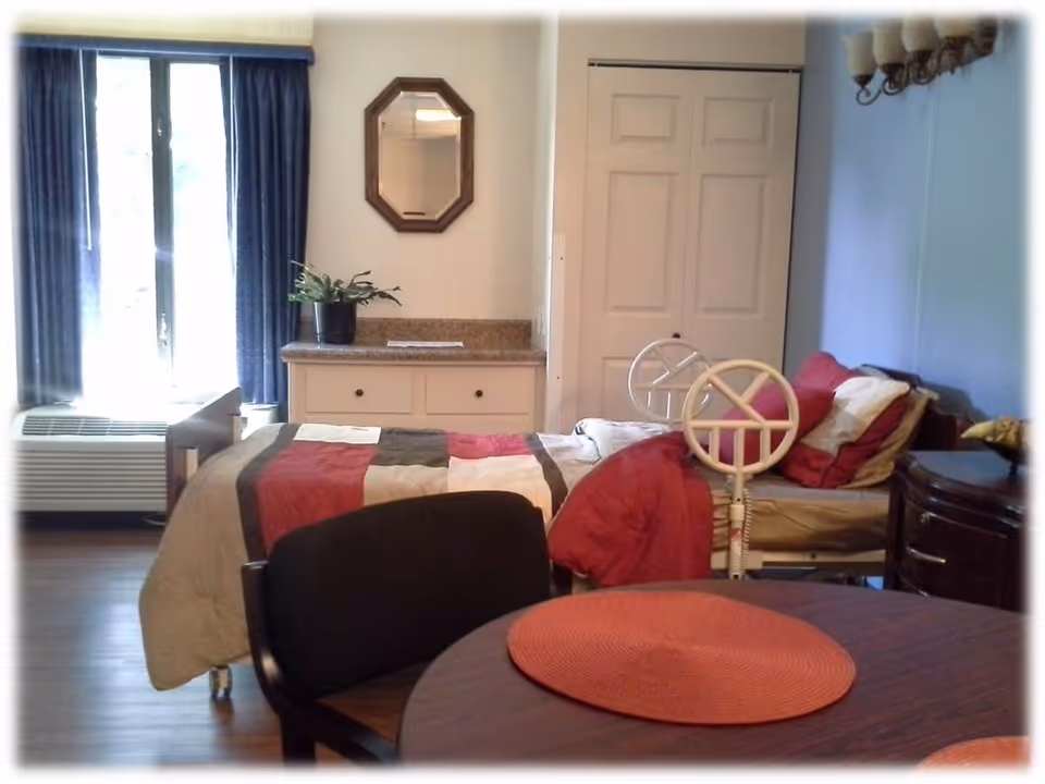 A senior living facility bedroom with a single bed covered in a multicolored quilt. The room has a window with blue curtains, a small dresser with a plant and a mirror above it, a round table with a red placemat, and a chair. There is a white closet door and a bedside table with a lamp.