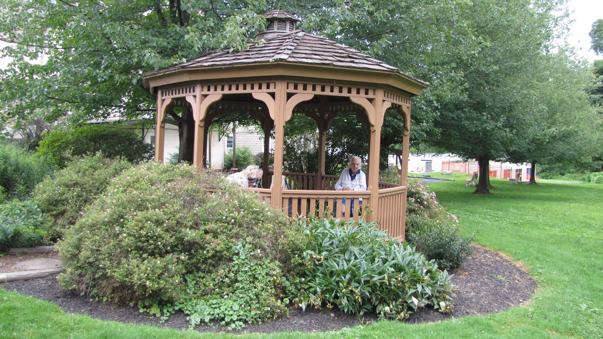 Two elderly women sitting inside a wooden gazebo surrounded by green bushes and trees in a garden area.