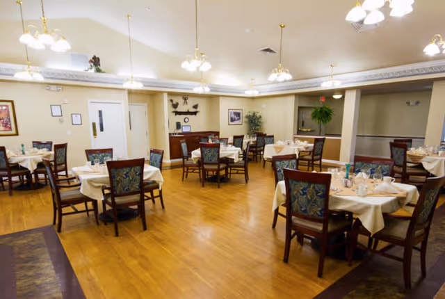 Spacious dining room with multiple round tables set with tablecloths and chairs beneath hanging chandeliers.
