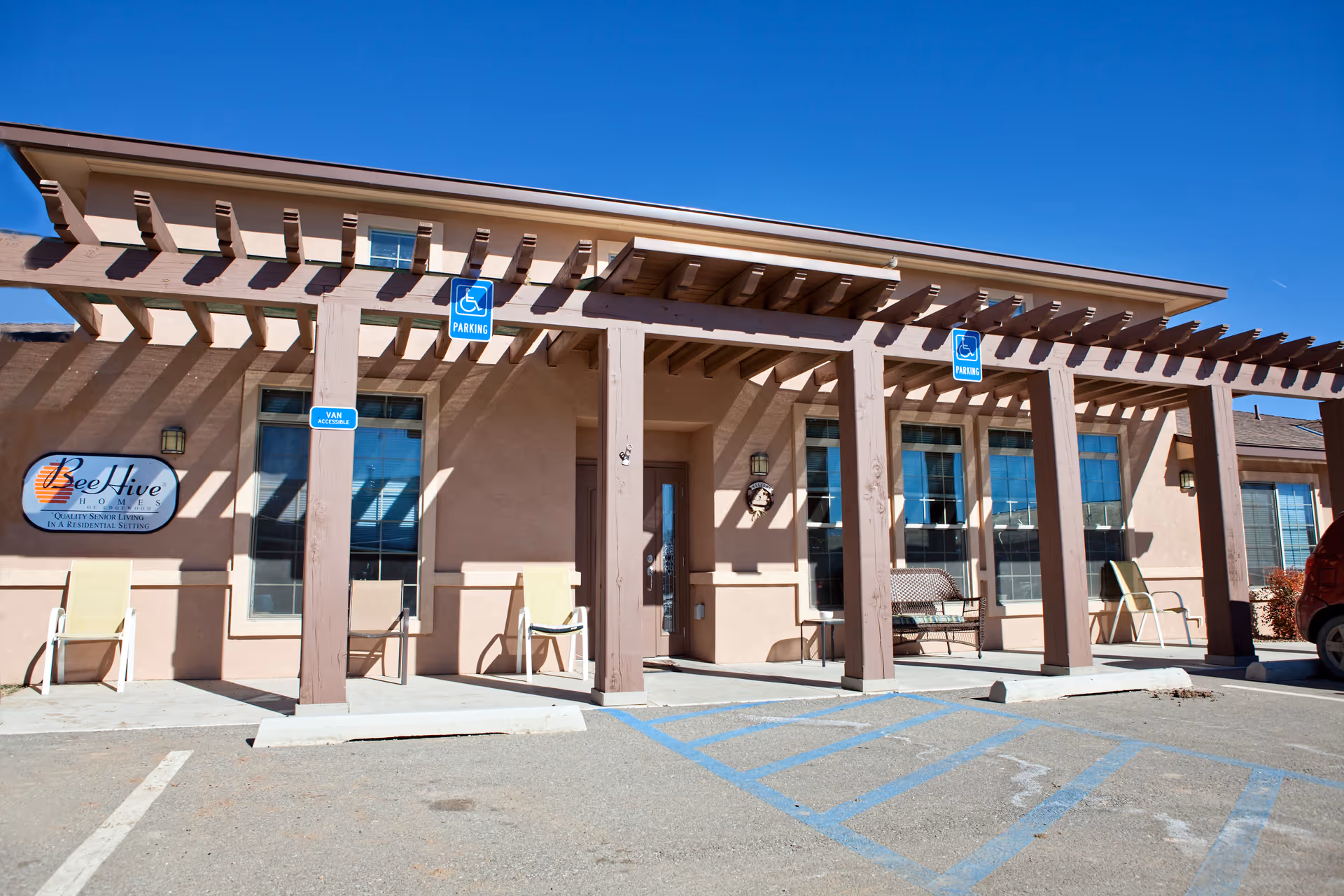 Exterior view of BeeHive Homes Assisted Living facility showing a covered entrance with wooden beams, several chairs, a bench, and handicap parking spaces in front under a clear blue sky.